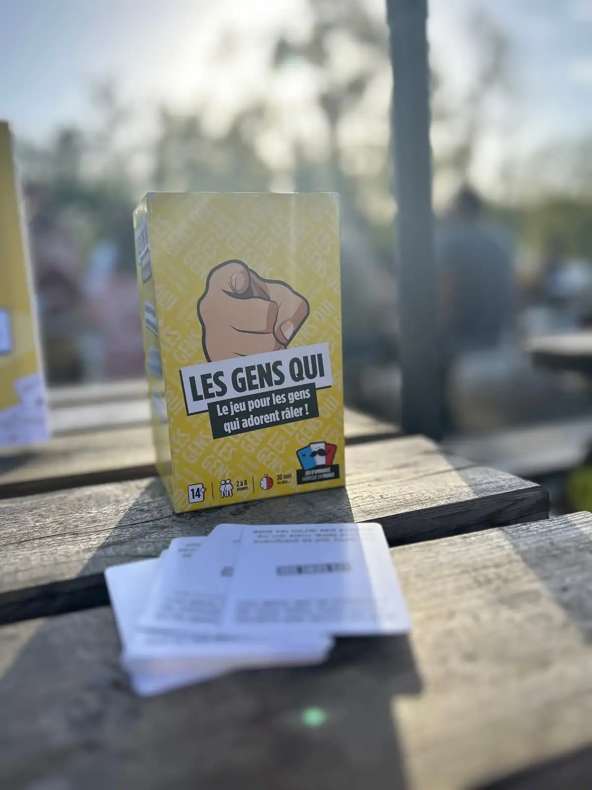 La boîte Les Gens Qui sur une table de terrasse en bois, cartes du jeu au premier plan, lumière de fin de journée
