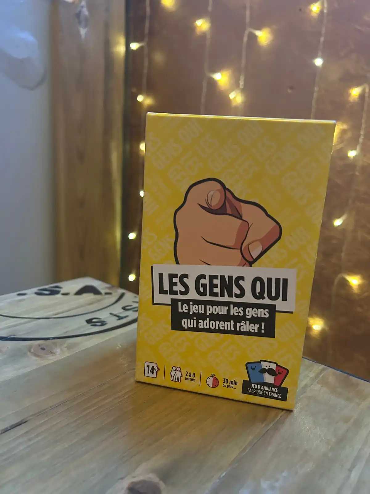 La boîte jaune Les Gens Qui posée sur une table en bois devant un rideau de guirlandes lumineuses, ambiance soirée