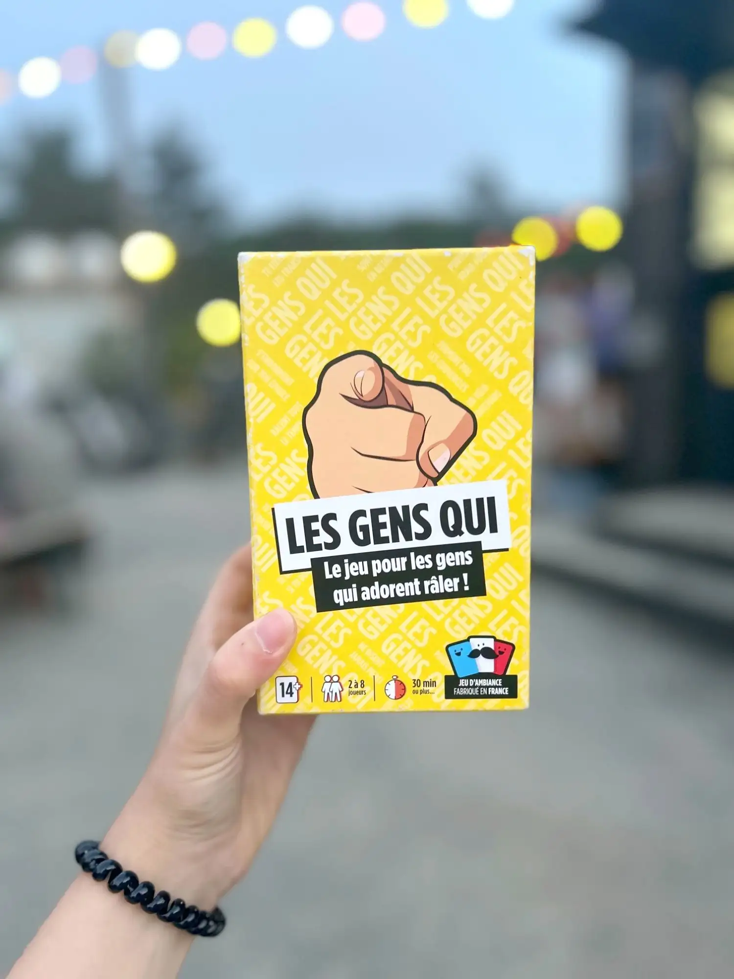 Une main brandit la boîte jaune du jeu Les Gens Qui, guirlandes lumineuses floues au crépuscule