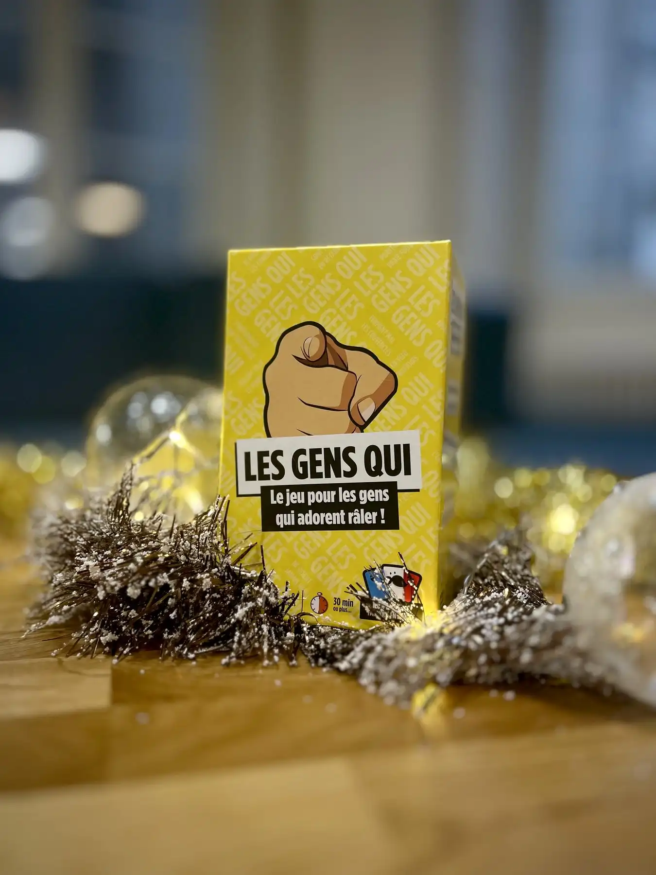 La boîte jaune Les Gens Qui posée sur une table en bois au milieu de guirlandes dorées, boules de Noël et lumières floues