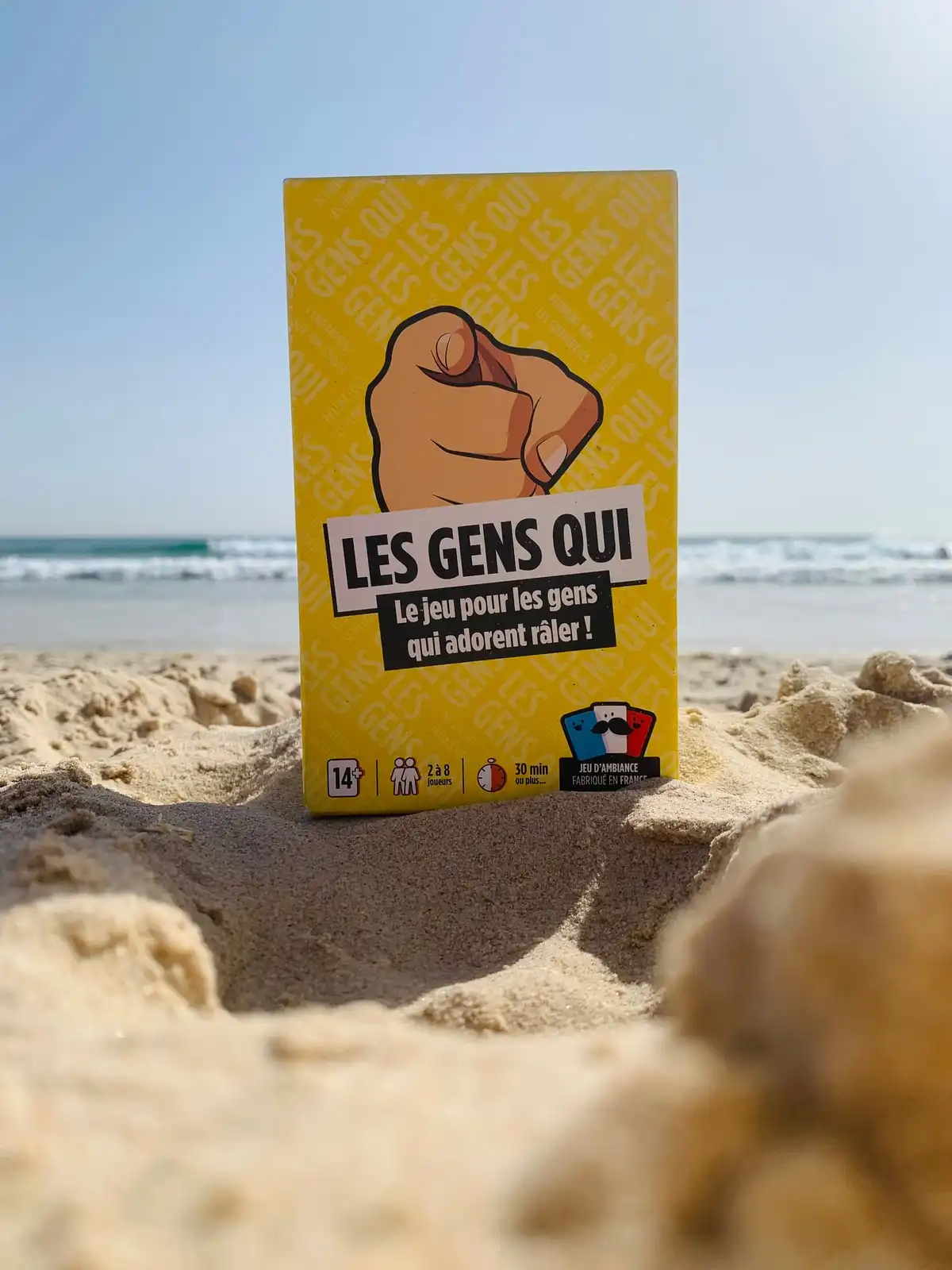 La boîte jaune du jeu Les Gens Qui plantée dans le sable d'une plage, la mer et le ciel bleu en arrière-plan