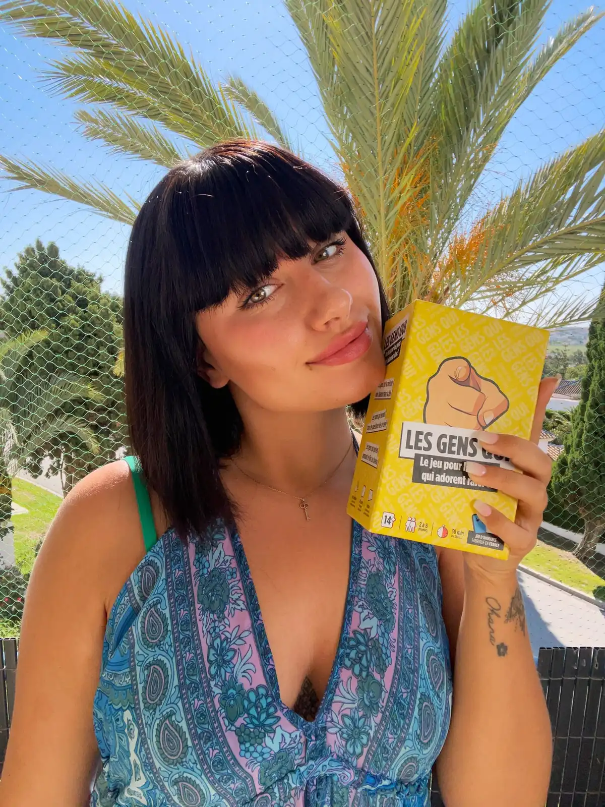 Une joueuse sourit, la boîte jaune Les Gens Qui serrée contre sa joue, sous un palmier en plein soleil