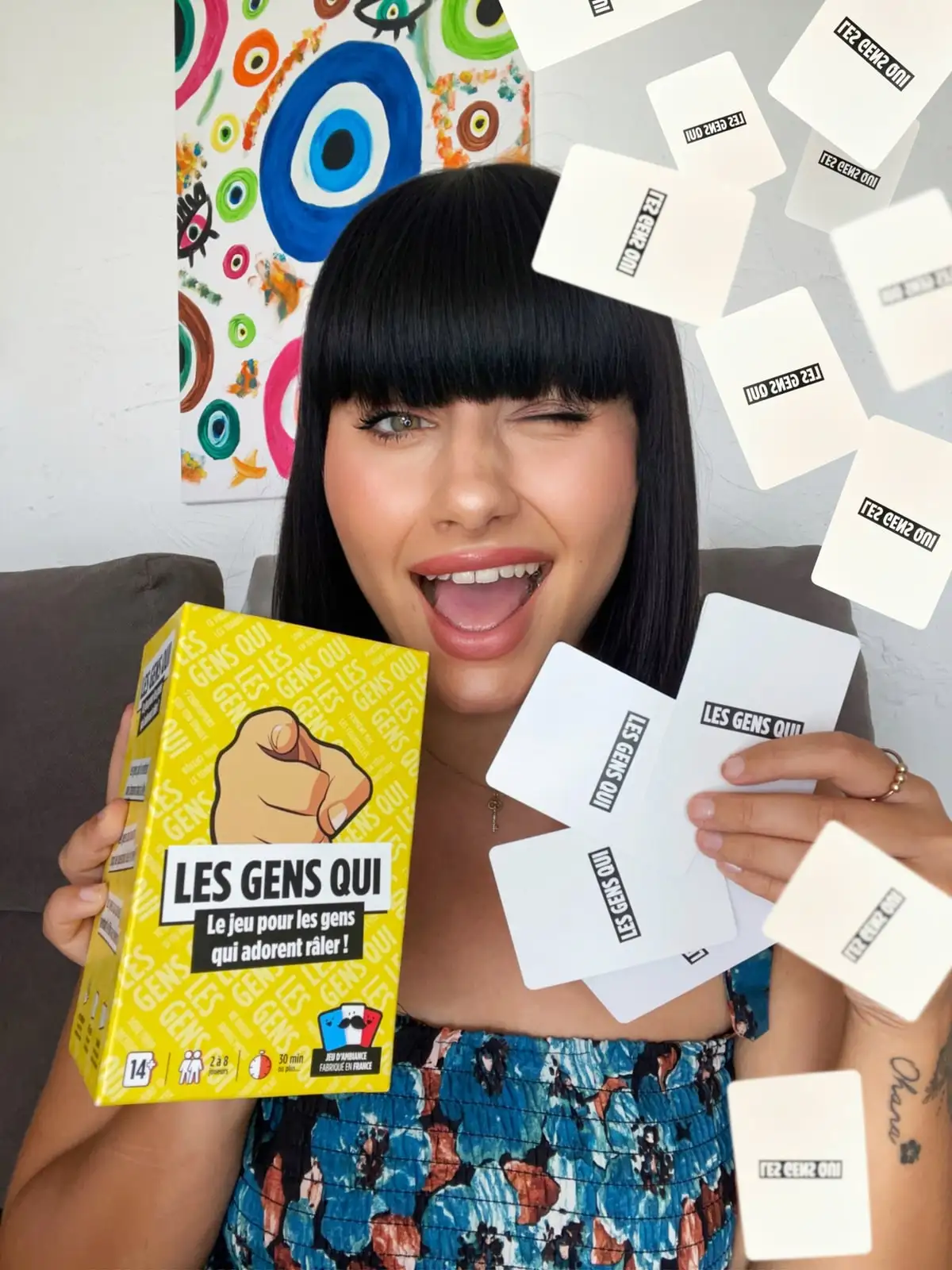 Une joueuse fait un clin d’œil, la boîte jaune Les Gens Qui à la main, pendant que des cartes volent autour d’elle