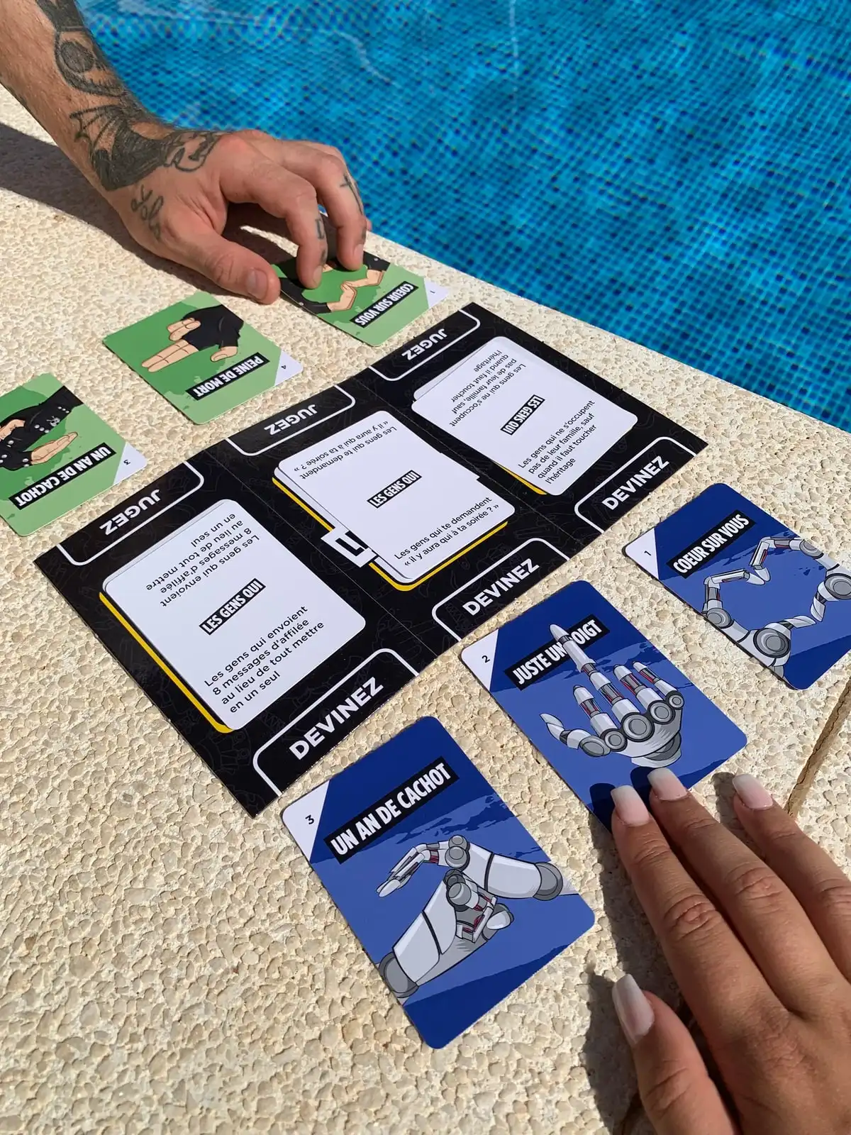 Au bord d’une piscine, des mains retournent les cartes Mains sous les cartes Les Gens Qui posées sur le plateau du Juge